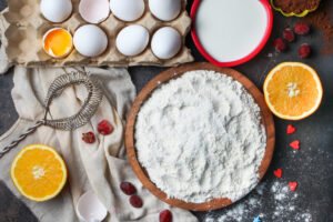 Flour with eggs, orange, milk, cocoa, whisk in a bowl on stone background, top view.